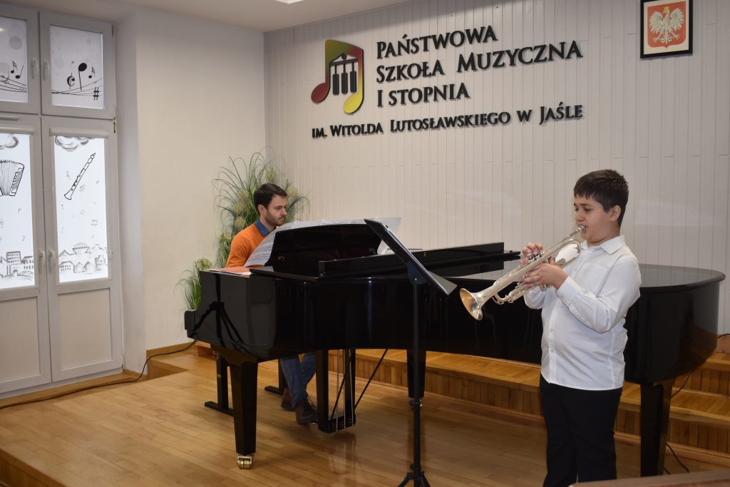 Na zdjęciu wykonanym w auli PSM widnieje uczeń grający na trąbce wraz z akompaniamentem fortepianu. Przy fortepianie nauczyciel Piotr Duda. Kolorystyka zdjęcia jest biało-czarno-brązowa. 