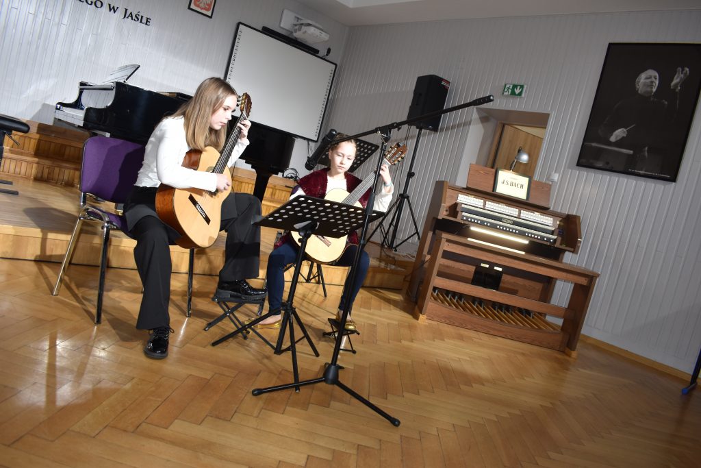 Na zdjęciu widnieje duet gitarowy złożony z uczennic szkoły. Zdjęcie wykonane jest w auli PSM. W tle fortepian oraz organy. Kolorystyka zdjęcia jest biało-brązowa.