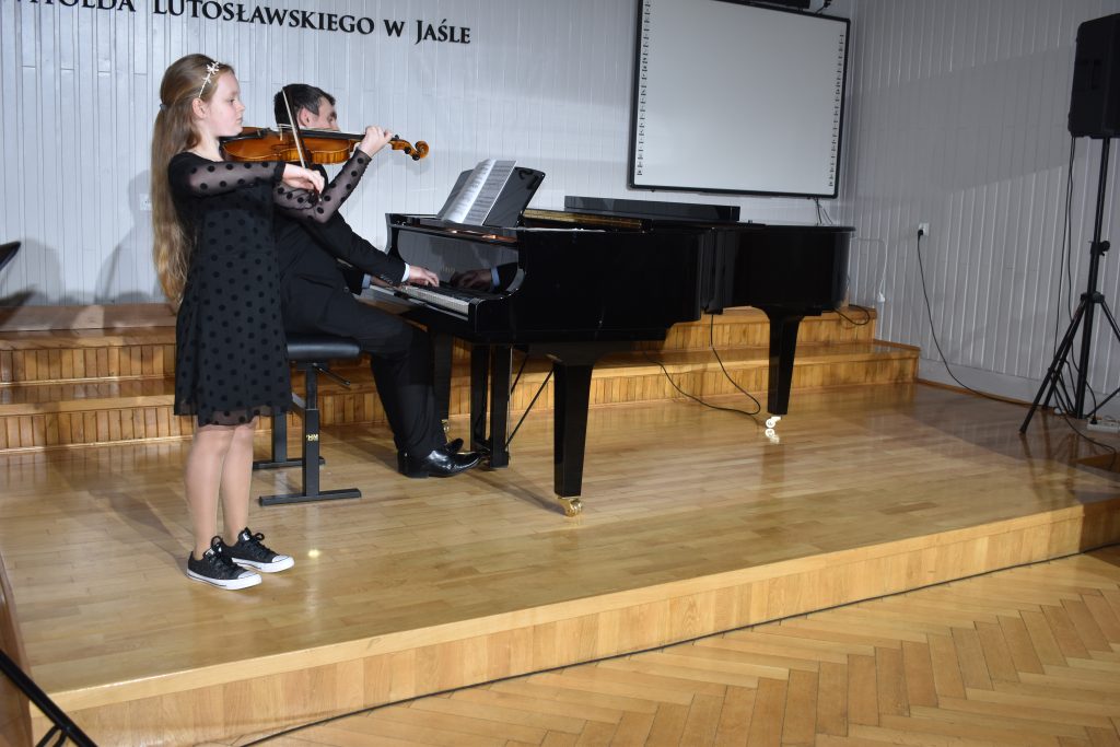 Na zdjęciu wykonanym w auli PSM widnieje uczennica grająca na skrzypcach z akompaniamentem fortepianu. Przy fortepianie nauczyciel Mateusz Gałuszka. Kolorystyka zdjęcia jest biało-czarno-brązowa.