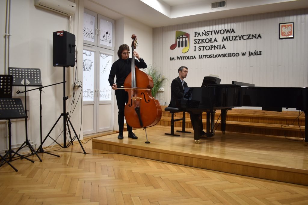 Na zdjęciu wykonanym w auli PSM widnieje uczeń grający na kontrabasie z akompaniamentem fortepianu. Przy fortepianie nauczyciel Mateusz Gałuszka. Kolorystyka zdjęcia jest biało-brązowa.